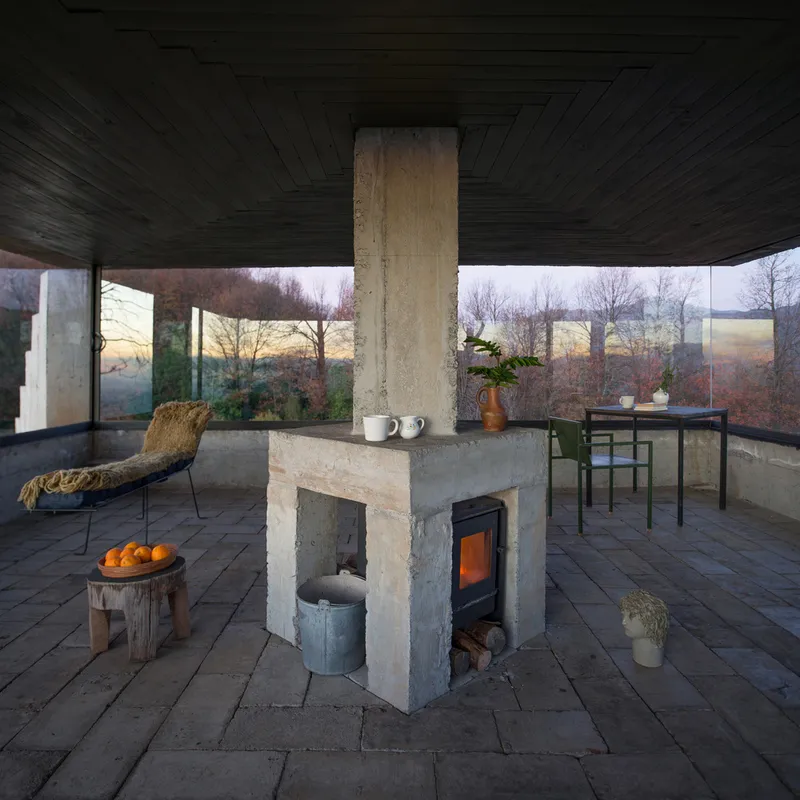Central fireplace with concrete chimney and stove surrounded by lounge chair and dining area at dusk