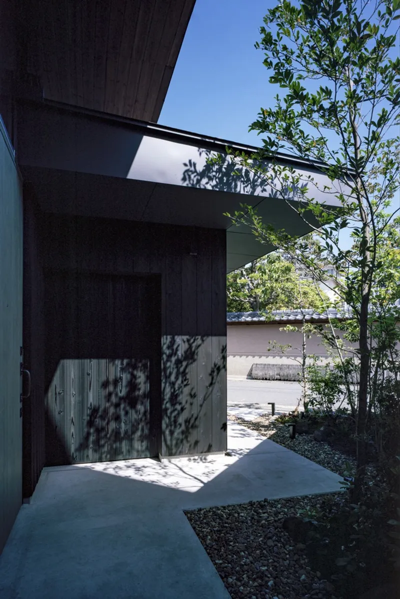 Covered entrance walkway with dark timber cladding and dappled tree shadows on the concrete path