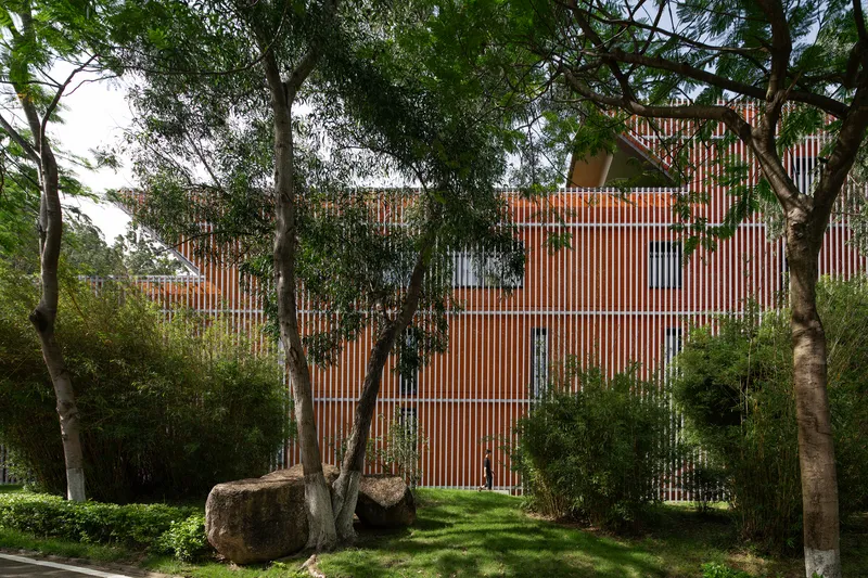 Vertical brick and glass screen facade seen through existing trees and boulders in landscaped garden
