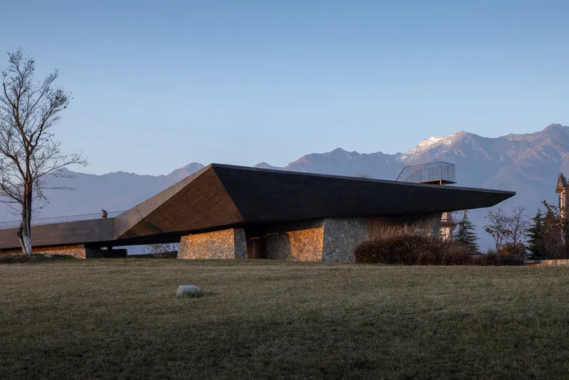 Angled black roof with timber underside over stone base walls at dusk with snow-capped mountains behind
