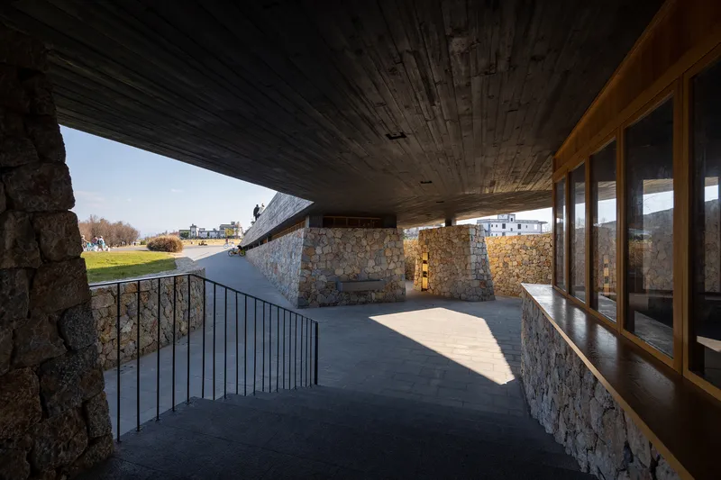 Covered terrace with charred timber ceiling, stone walls, and timber-framed glazing overlooking open fields