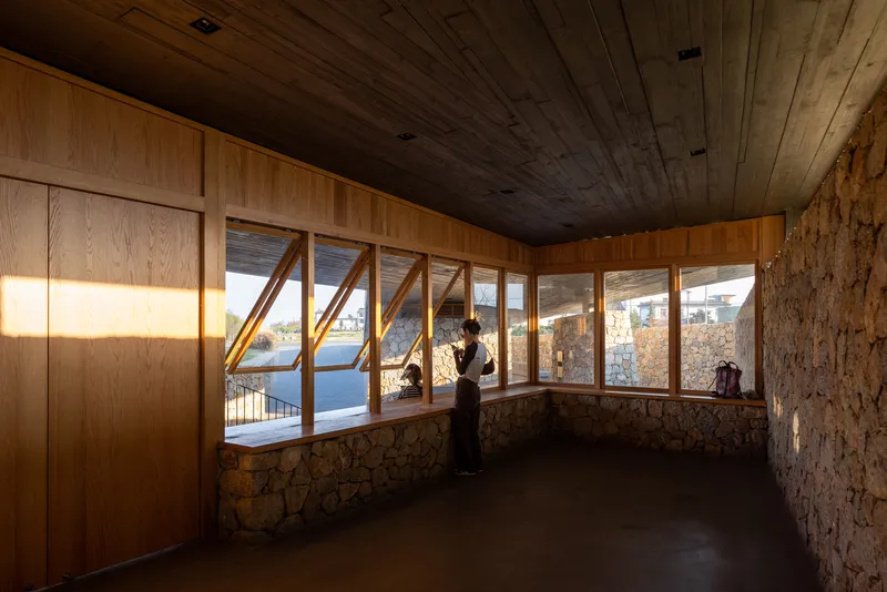 Interior room with angled timber-framed windows, stone wall base, and a figure standing at the glass