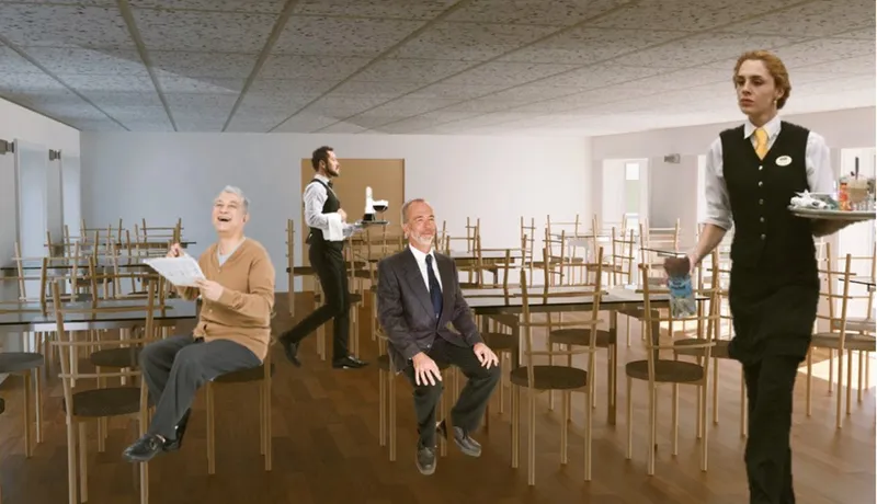Dining hall with suspended ceiling tiles and light wood chairs arranged in rows with staff and residents