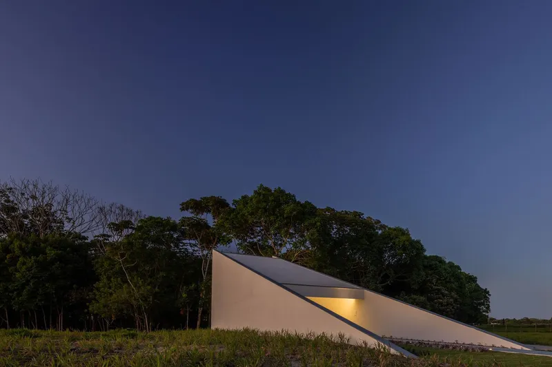 Angled roof plane with illuminated slit window photographed at dusk against the treeline