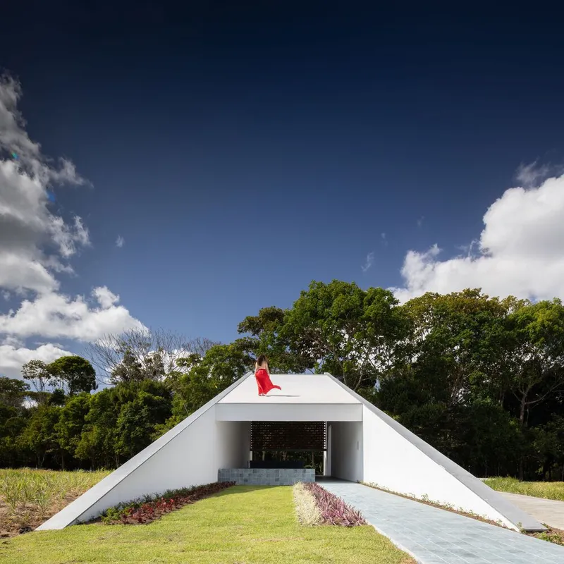 White pyramidal roof rising from planted lawn with red flag at apex under blue sky