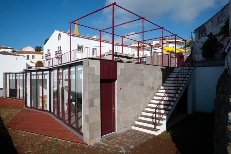Concrete block volume with glazed walls and red steel rooftop structure under blue sky