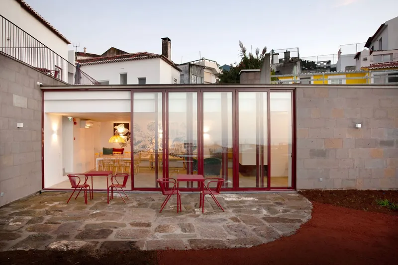 Glass pavilion with maroon steel frames opening onto a cobblestone patio at dusk
