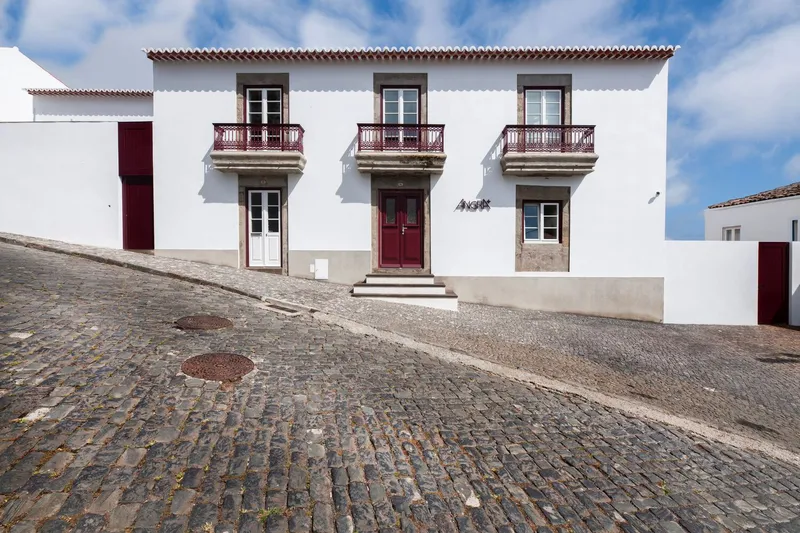 White rendered facade with three balconies and maroon doors facing cobbled street