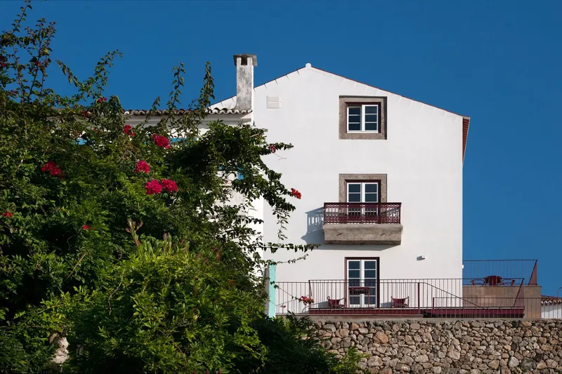 White stucco facade with framed windows and iron balconies behind flowering bougainvillea and a stone retaining wall