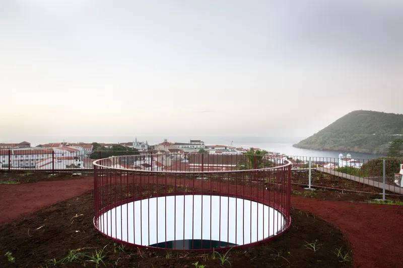 Circular red steel railing enclosing a planted courtyard overlooking a harbor and hillside at dusk