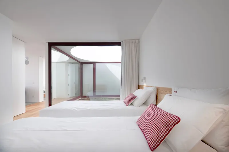 Bedroom with light wood flooring and red-framed curved window opening to a terrace courtyard