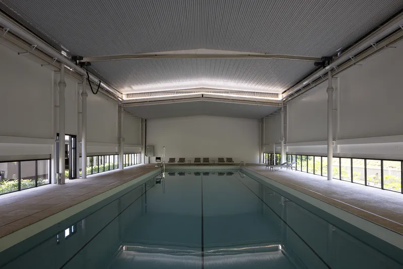 Indoor lap pool with white roller shades drawn over floor-to-ceiling windows beneath a corrugated metal ceiling