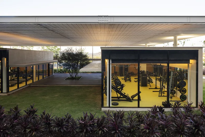 Glass-walled fitness pavilion with dark purple foliage bed and a glimpsed courtyard beyond at dusk