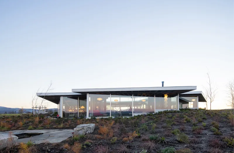 Glass-walled pavilion with flat overhanging roof nestled in a rocky hillside landscape at dusk