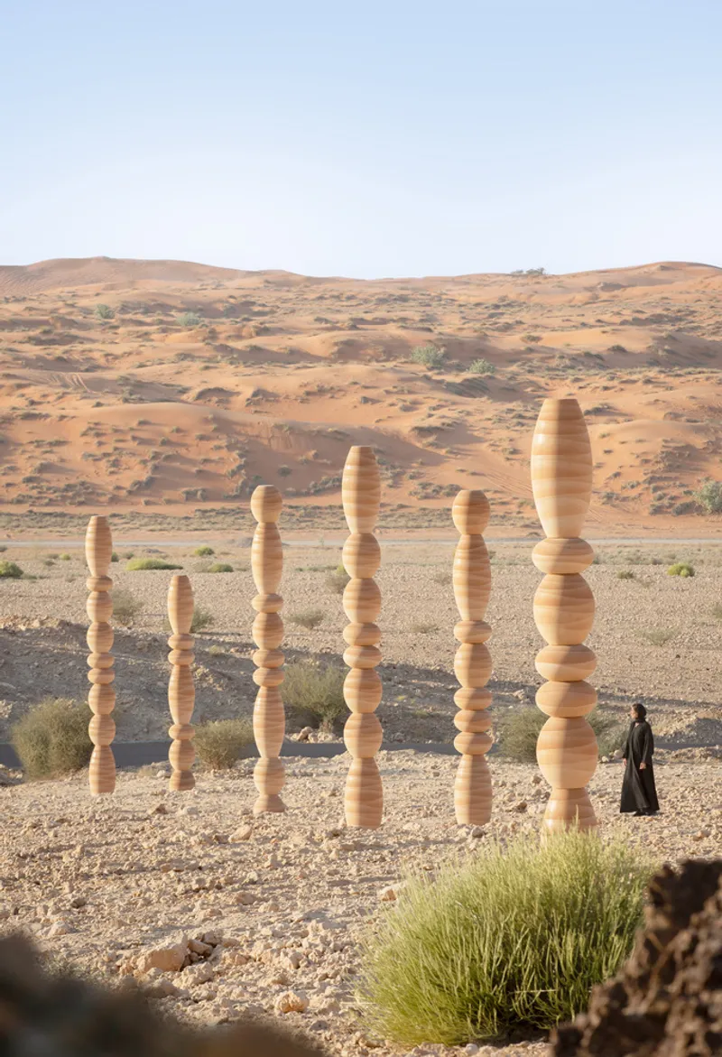 Seven varying-height timber totems standing in gravel with a person walking and sand dunes beyond