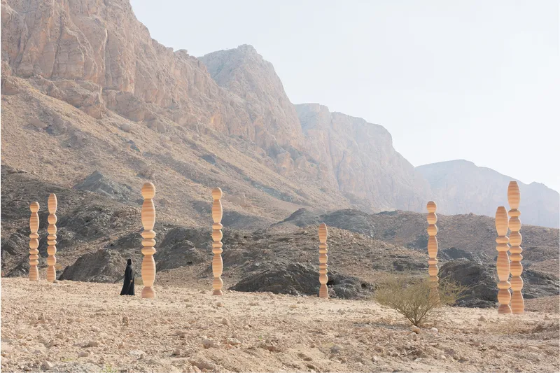 Figure in black walking past the timber columns on the stony plateau beneath towering cliffs