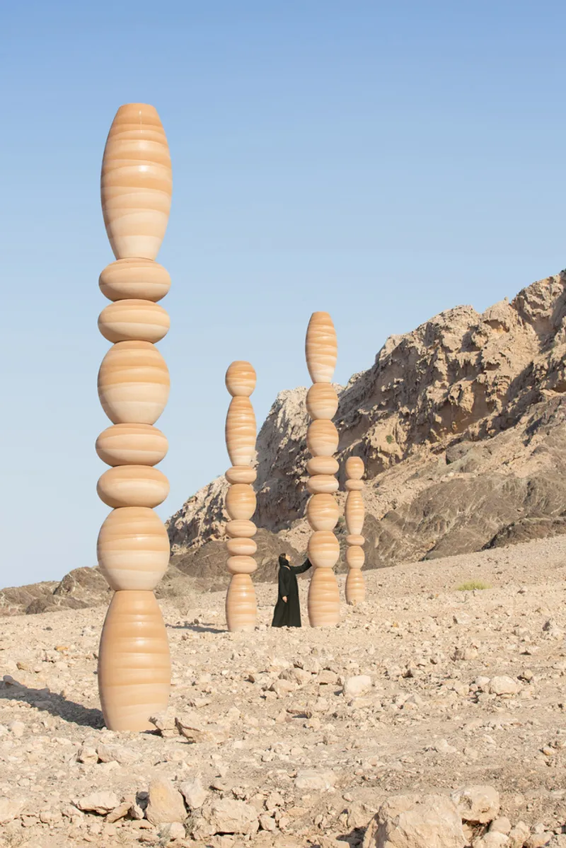 Three laminated wood columns of varying heights with a figure walking between them near rocky hills