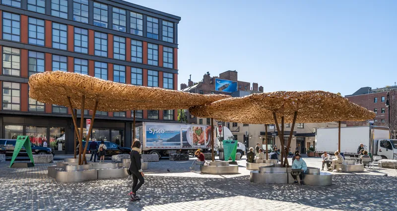 Wide view of clustered woven timber shade structures on a paved plaza with people seated at tables below