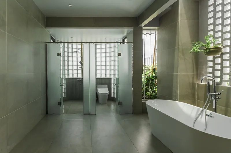 Grey-tiled bathroom with freestanding tub and glass-enclosed shower stalls facing perforated concrete block screens