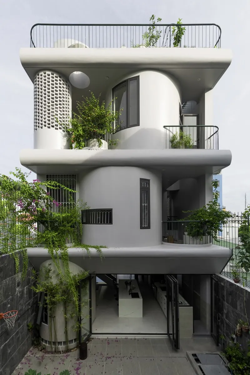 Three-story white facade with rounded balconies, cascading plants, and glass block cylinder framed by dark retaining walls