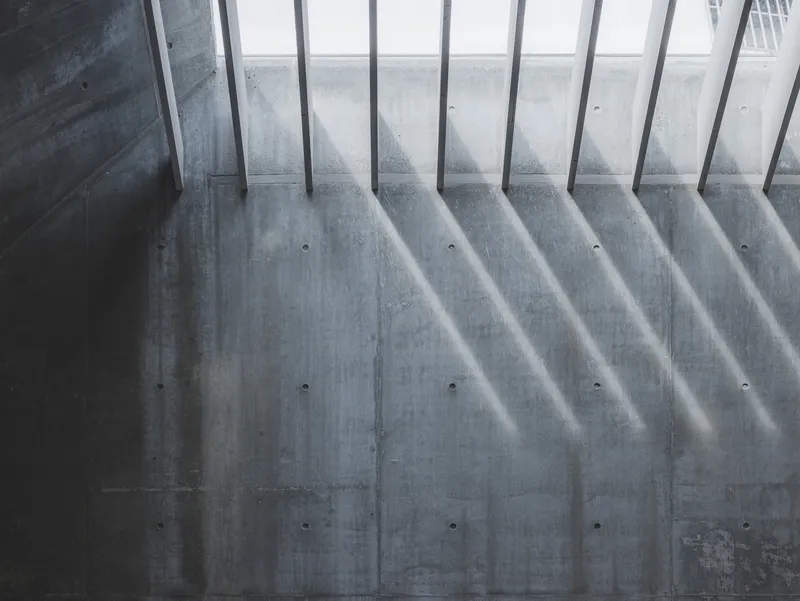 Board-formed concrete wall with diagonal shadows cast by vertical steel railings above