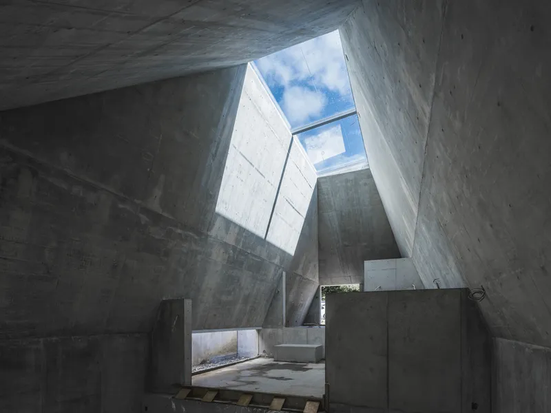 Upward view through a cast concrete atrium with angular skylights framing blue sky and clouds