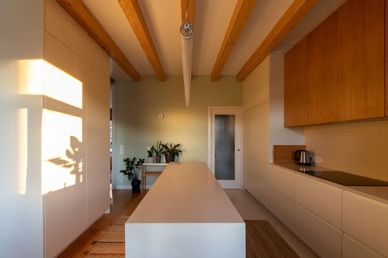 Kitchen with white island bench and timber cabinetry beneath exposed ceiling beams in warm light