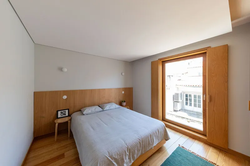 Bedroom with timber-framed window overlooking neighboring rooftops under diffused daylight
