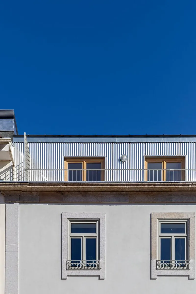 Street facade showing a corrugated metal rooftop extension above a white plastered base with framed windows under blue sky