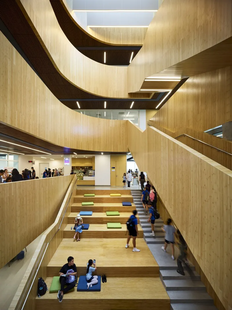 Timber-clad atrium with curved balconies and stepped seating where students gather below skylights