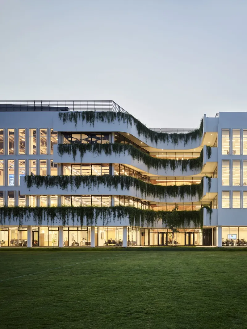 Facade with stacked terraces draped in cascading greenery above a grass field at twilight
