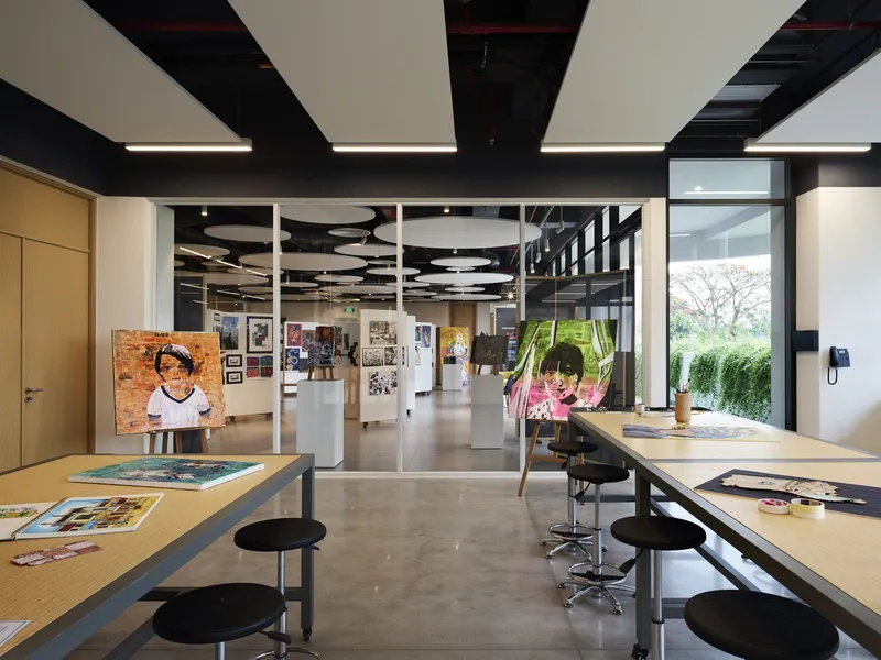Art studio with worktables, black acoustic ceiling panels, and student paintings visible through interior glazing