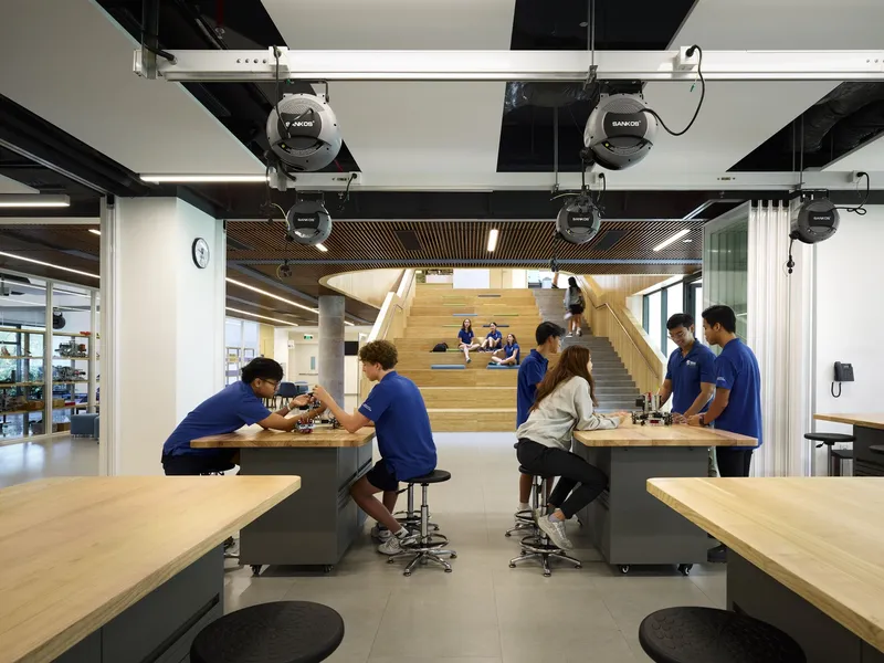 Workshop space with exposed ceiling infrastructure and students working at movable benches beneath timber stairs