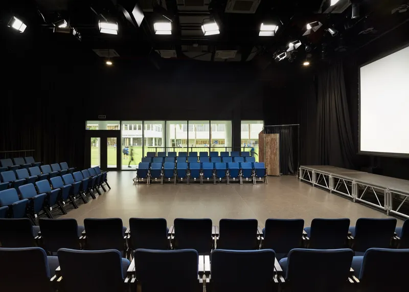 Auditorium interior with black ceiling grid, blue seats, and glazed doors overlooking courtyard