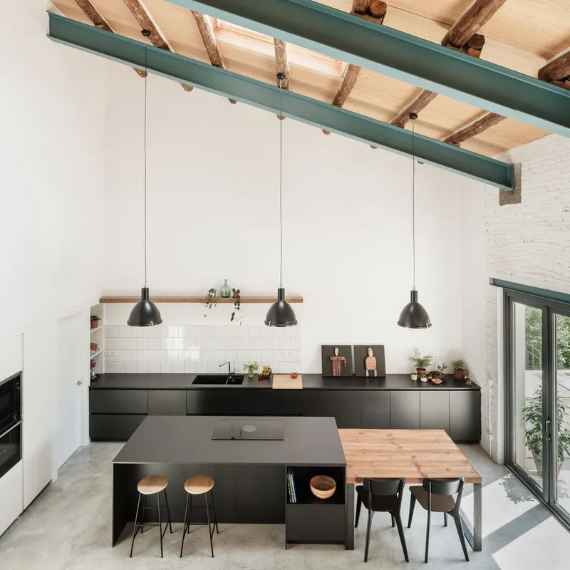 Kitchen with black island and timber dining table beneath three pendant lights and exposed rafters