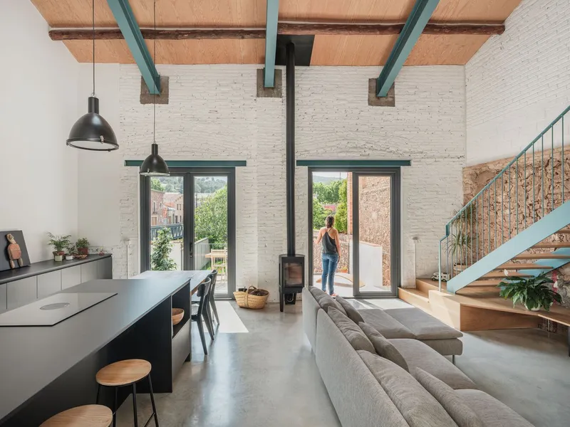 Double-height living space with painted white brick walls, exposed timber ceiling with green steel beams, and woman standing in doorway