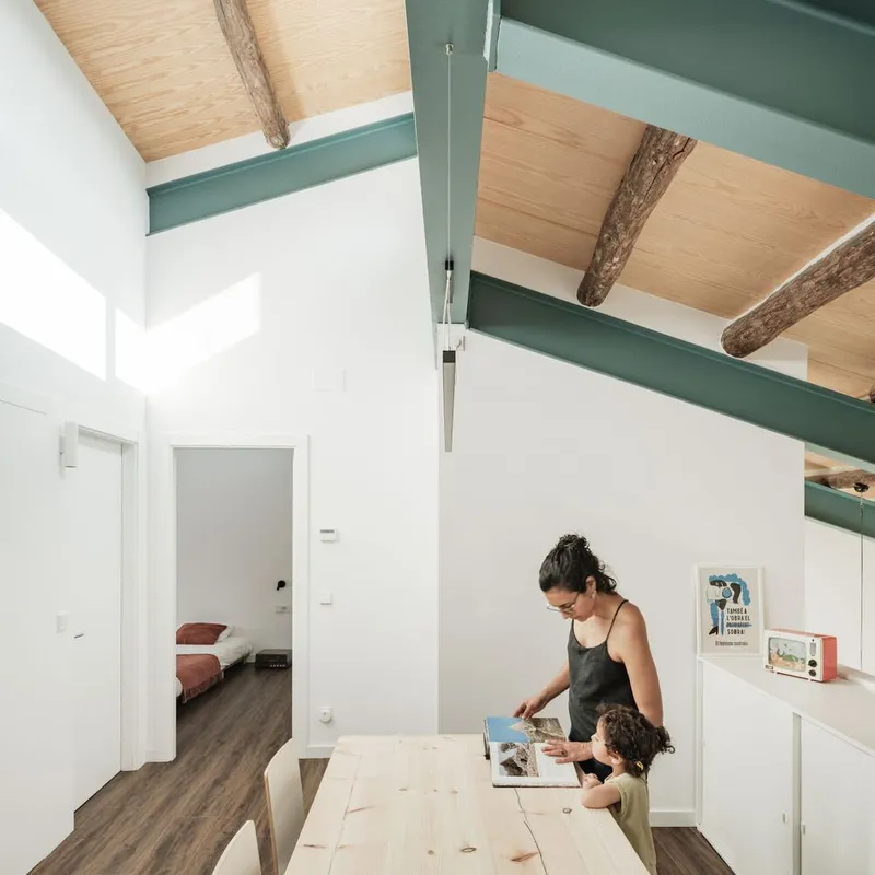 Vaulted ceiling with exposed timber beams and green steel trusses above a pale timber dining table with adult and child