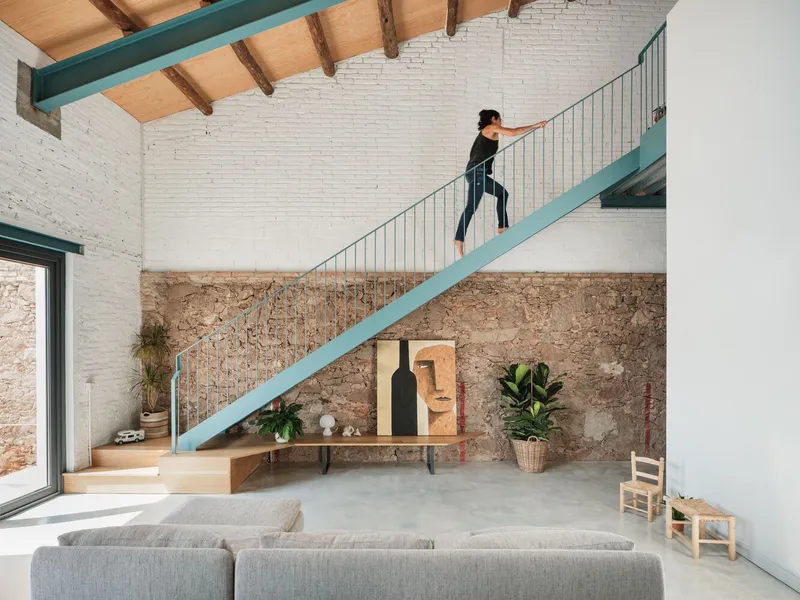 Double-height living room with turquoise steel staircase ascending past exposed stone wall and painted white brick