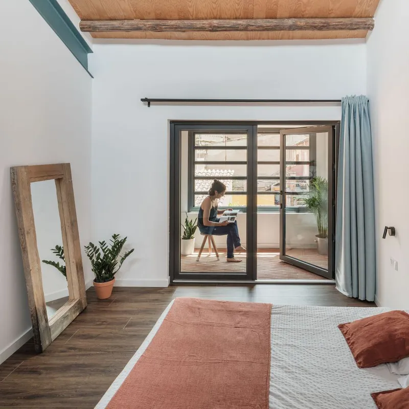 Bedroom with glazed doors opening to a terrace where a person sits reading in sunlight