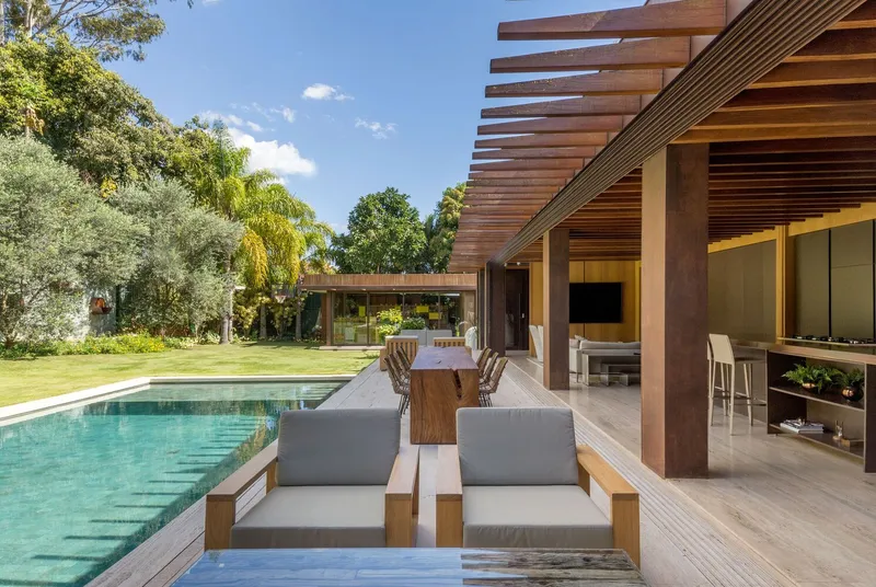 Poolside lounge seating beneath timber pergola with lawn and trees in afternoon sun