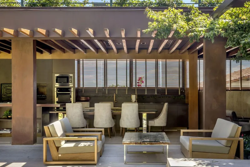 Open-air living pavilion with timber pergola structure, louvered screens, and kitchen bar viewed through dappled sunlight
