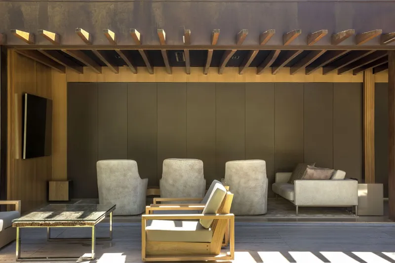 Covered terrace with exposed timber rafters and vertical panel wall behind upholstered seating in afternoon light