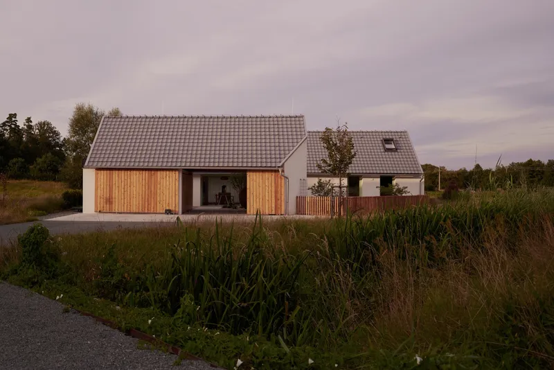 Two gabled volumes with vertical timber cladding and white render set in tall grasses at dusk