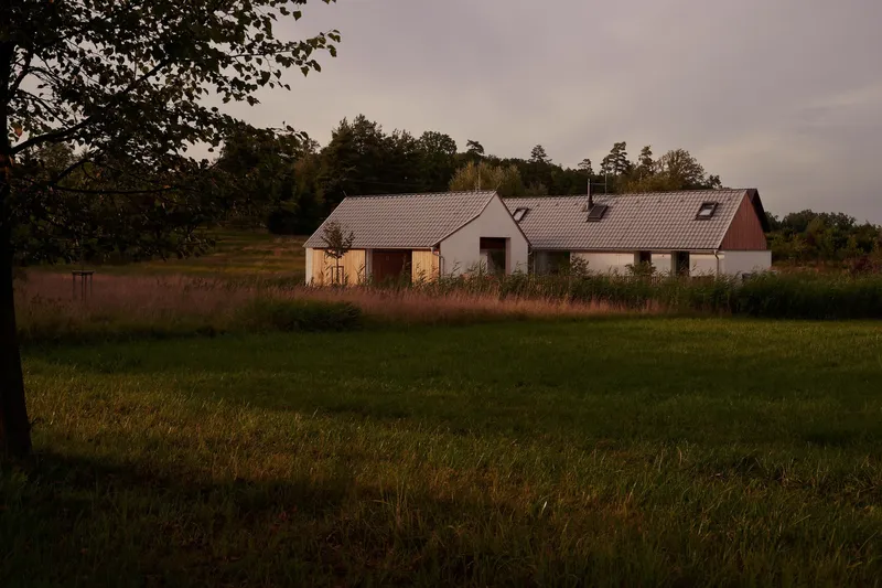 Linked gabled buildings with tile roofs and timber cladding viewed across a meadow in evening light