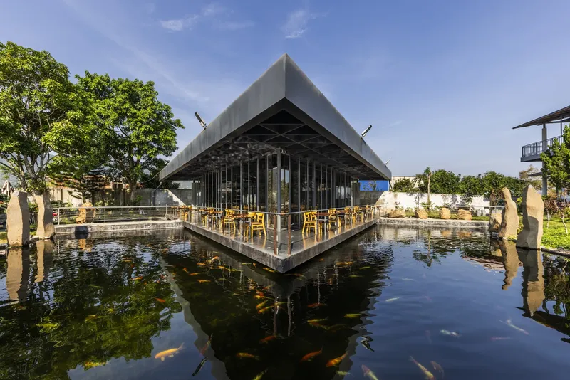 Triangular pavilion with full-height glass walls and exposed steel ceiling structure surrounded by reflecting pond