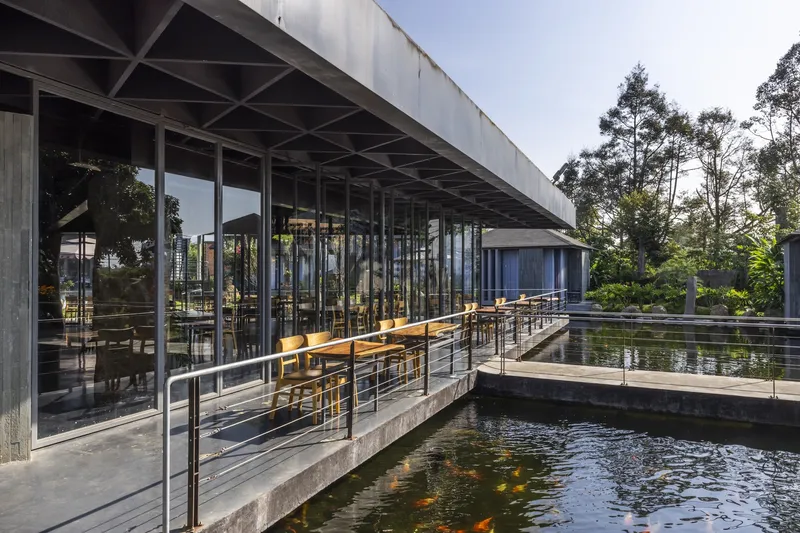 Covered terrace with slim steel columns and concrete walkway spanning koi ponds with trees in the background