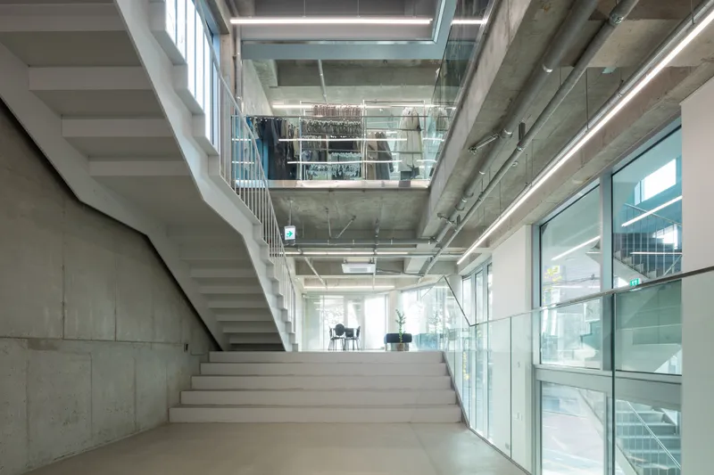 White staircase ascending through a multi-story atrium with glass balustrades and exposed concrete structure