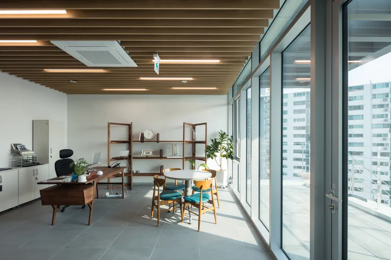 Office space with wood-slat ceiling, full-height glazing opening to a terrace, and midcentury furnishings in afternoon light