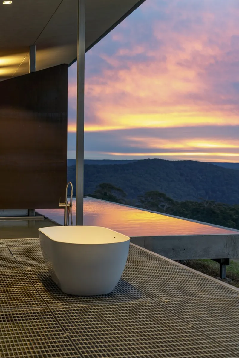 Freestanding bathtub on a metal grate floor with full-height glazing overlooking the infinity pool at sunset
