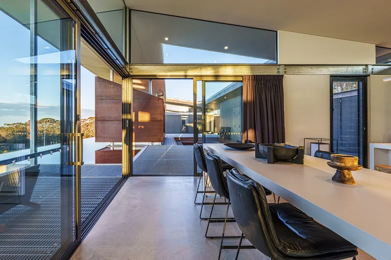 Dining area with white table extending toward glass doors opening onto metal grate deck at sunset